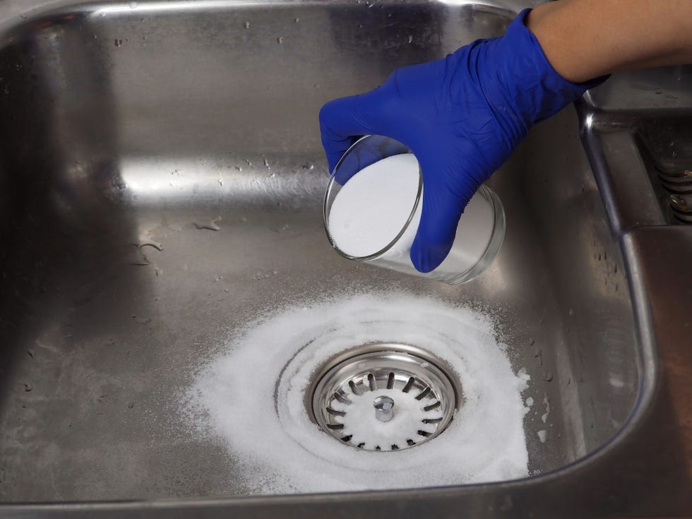 garbage disposal cleaner, gloved hand putting baking soda on drain in kitchen sink